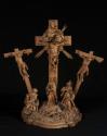 The image shows a sculptural representation depicting a crucifixion scene. In the center, there is a tall crucifix with a figure nailed to the cross. On both sides of the central cross, there are additional figures also hanging from crosses. Beneath the crucifix, a group of figures is arranged, including a central figure that represents a female figure, with two more figures standing on either side. The entire composition is richly decorated and conveys a religious theme.

[AI-generated with C…