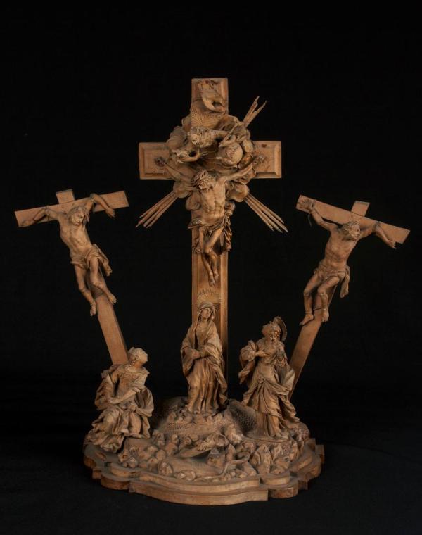 The image shows a sculptural representation depicting a crucifixion scene. In the center, there is a tall crucifix with a figure nailed to the cross. On both sides of the central cross, there are additional figures also hanging from crosses. Beneath the crucifix, a group of figures is arranged, including a central figure that represents a female figure, with two more figures standing on either side. The entire composition is richly decorated and conveys a religious theme.

[AI-generated with C…